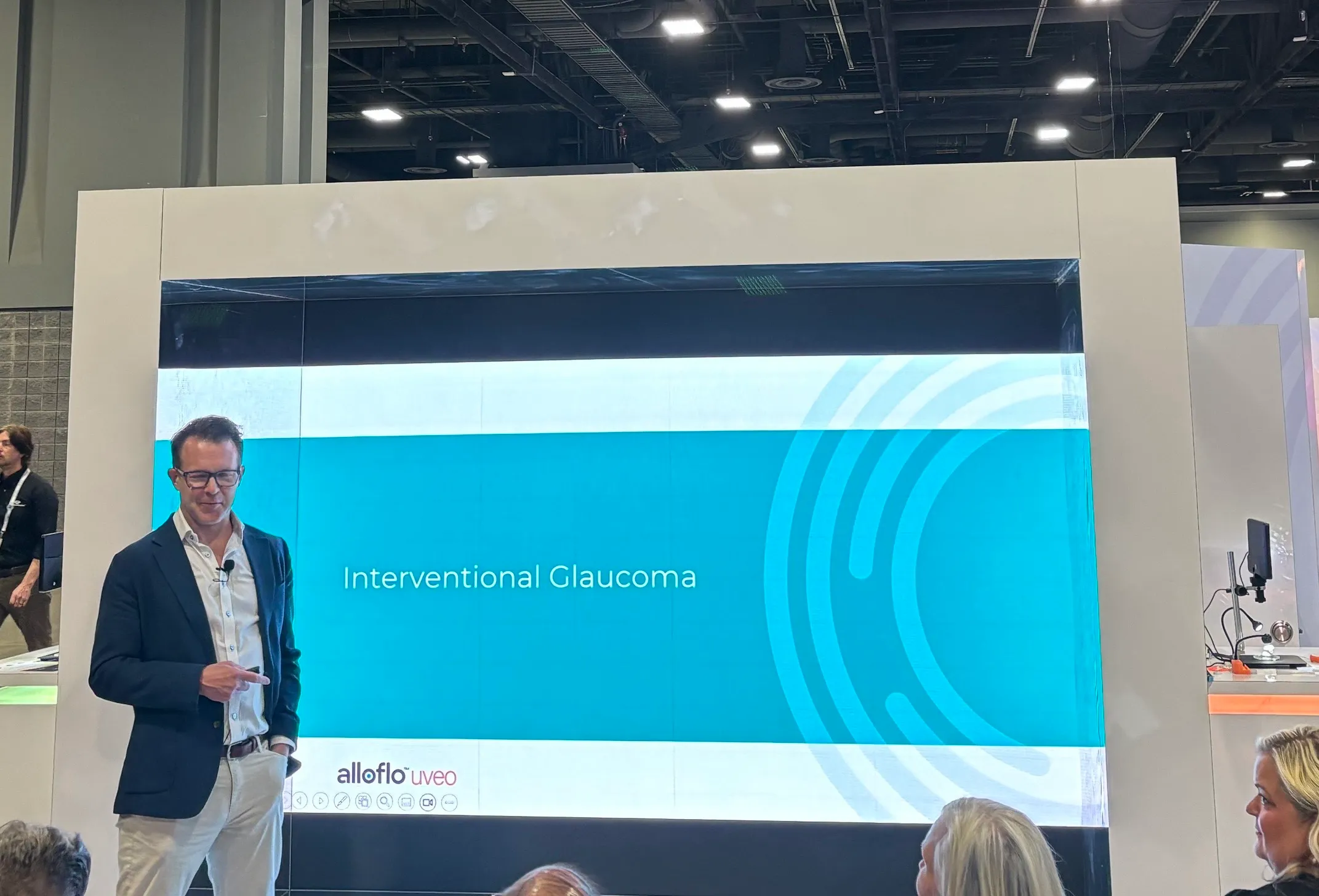 A speaker presenting at an AlloFlo Uveo booth talk in front of a screen reading "Interventional Glaucoma"
